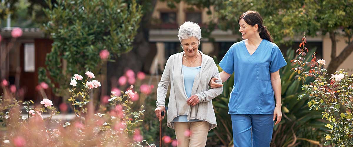 A health care professional walks arm in arm with a senior woman outside through a garden