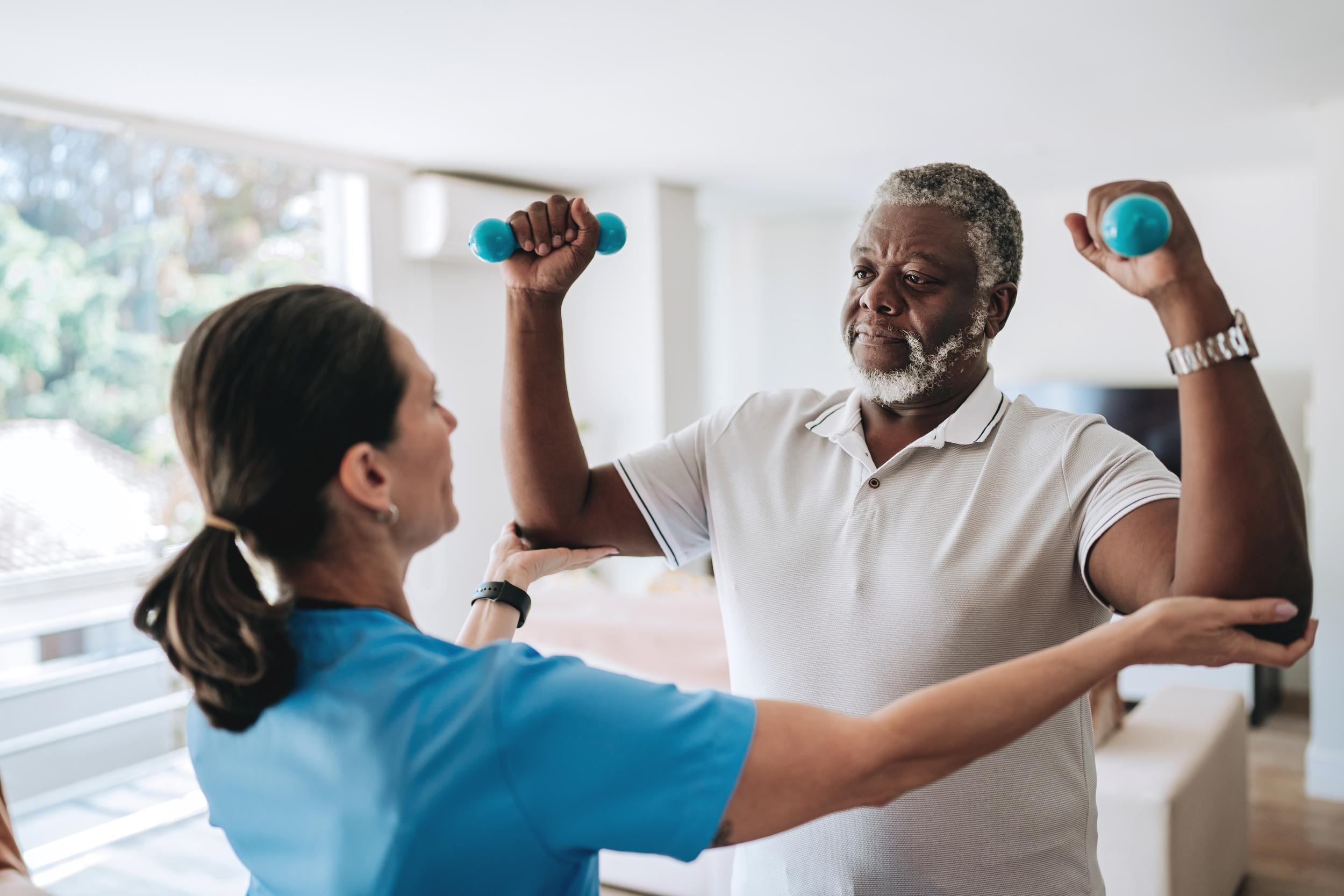 Senior man holding up dumbbells while female healthcare worker is assisting